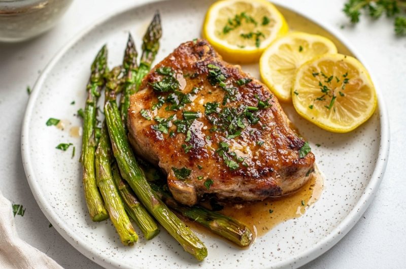 Succulent pork chops topped with a luscious lemon butter sauce, served with a side of asparagus in a plate.