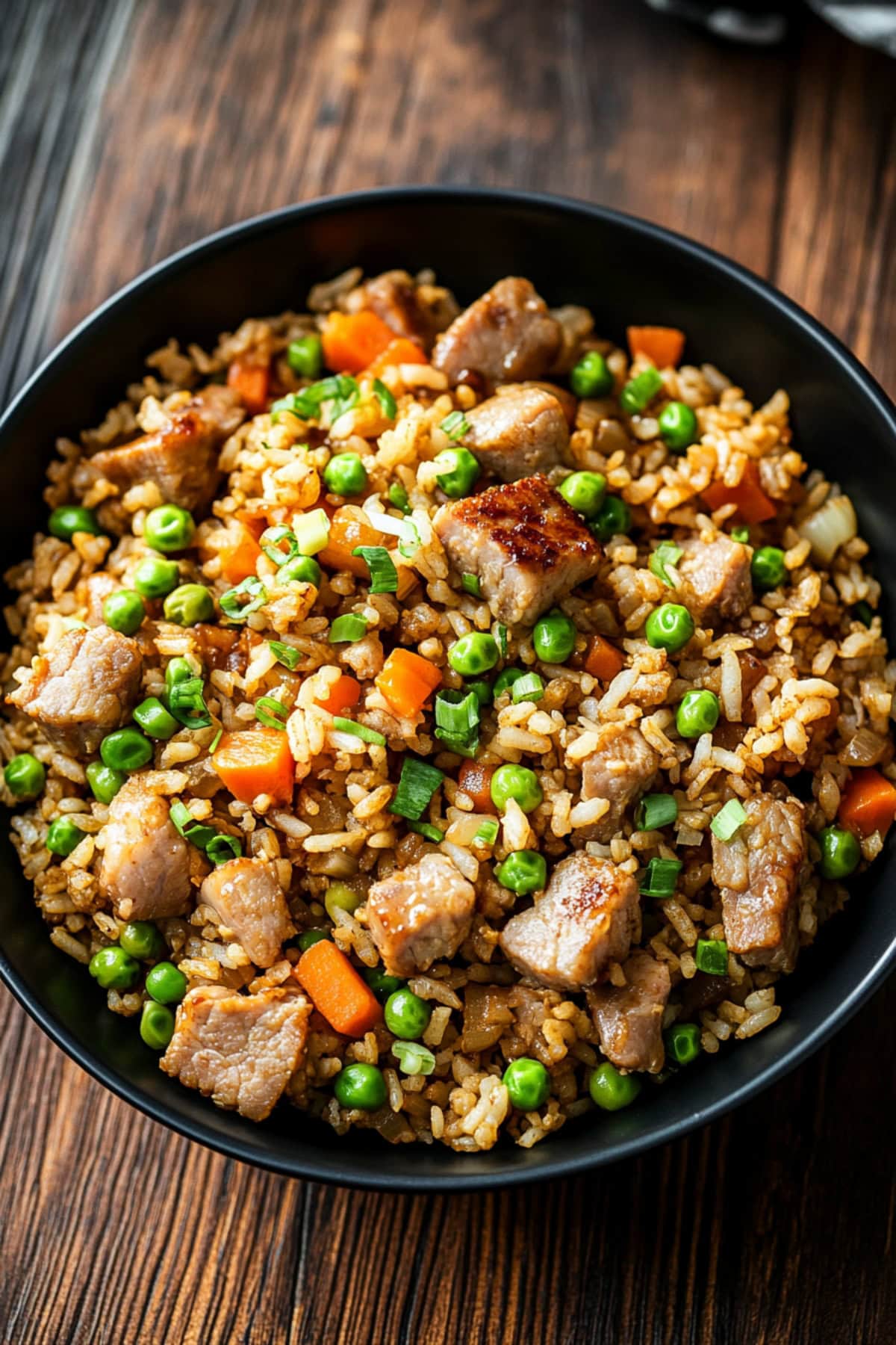 A black bowl of pork fried rice with carrots, peas and onions.