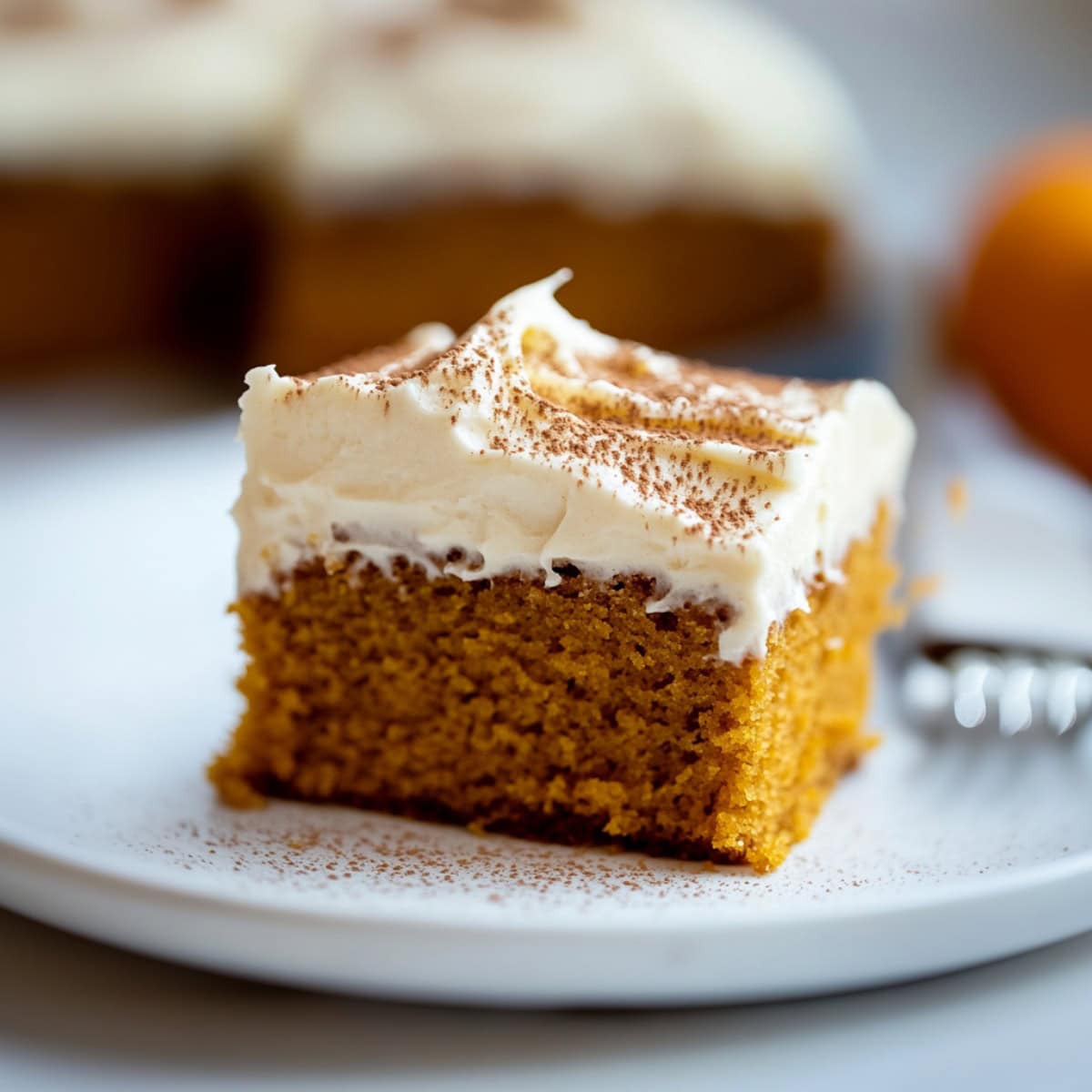 A slice of pumpkin cake with cream cheese frosting, sprinkled with ground cinnamon.