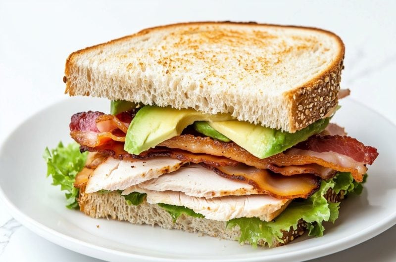 Sliced in half serving of turkey avocado sandwich served in a white plate.