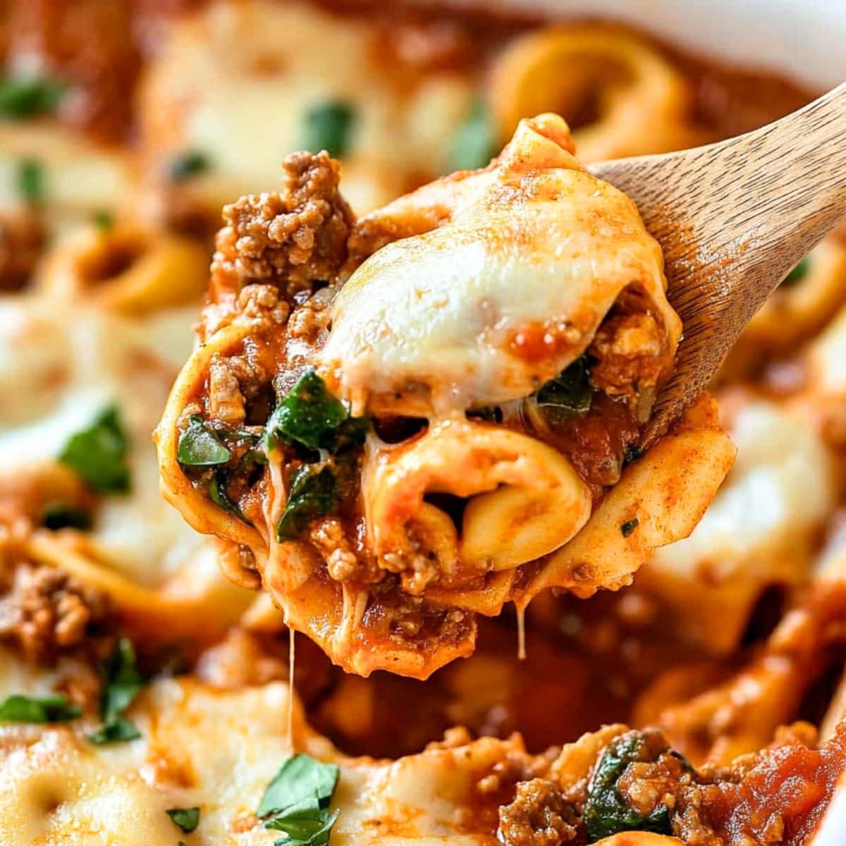 Cheesy baked tortellini with ground beef in cheesy marinara sauce scooped with a wooden spoon.