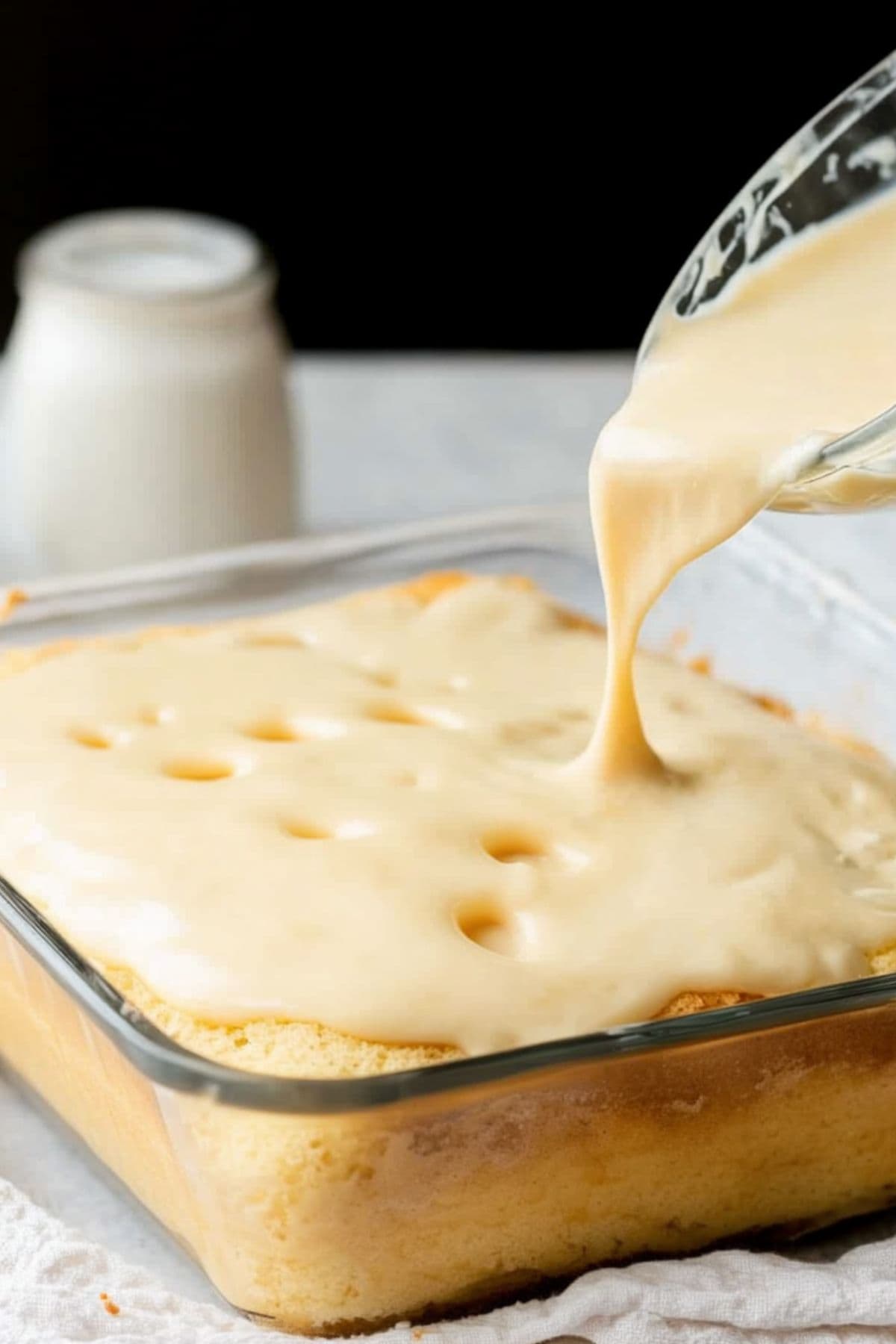 Vanilla pudding being poured over a vanilla cake in a glass baking dish