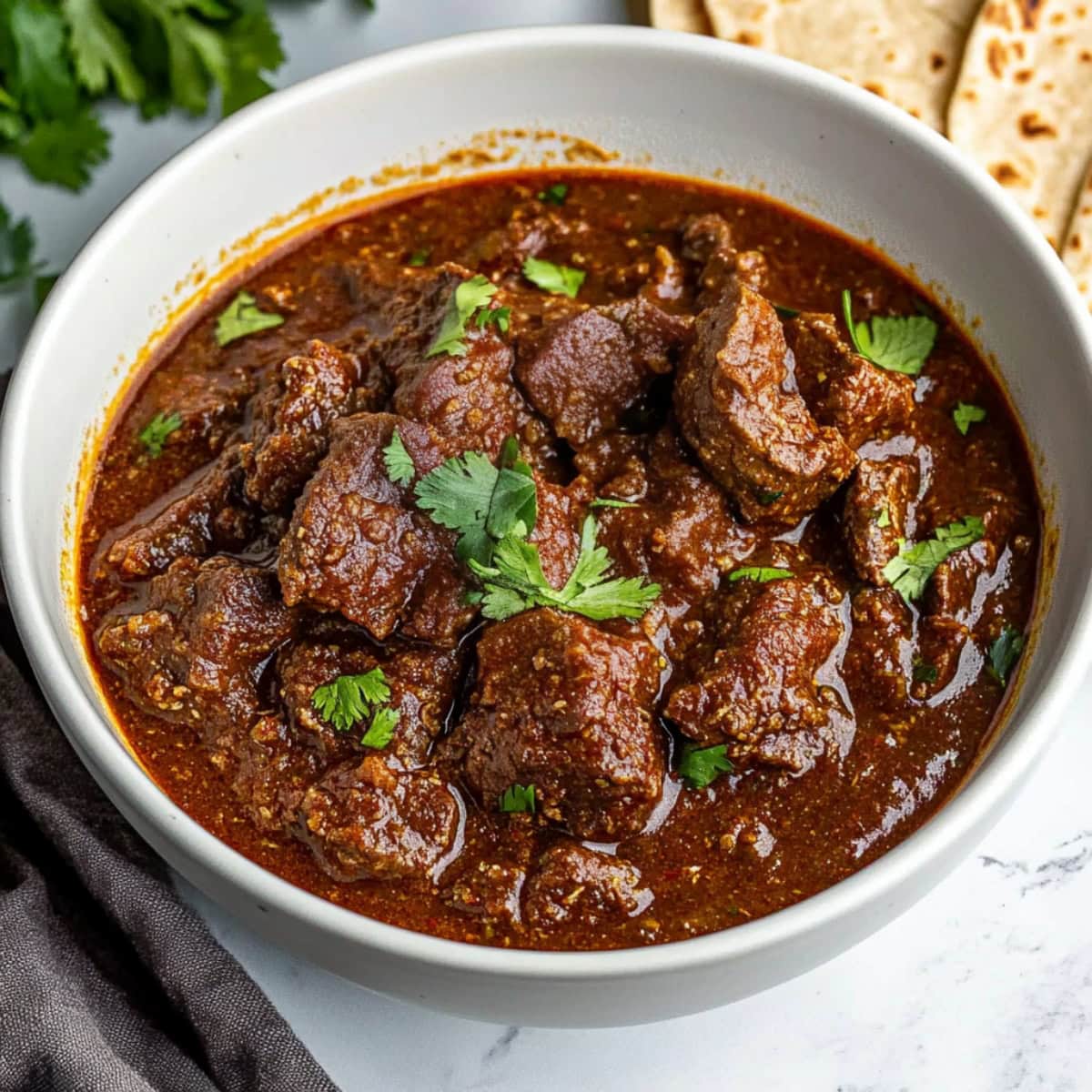 A bowl of chili Colorado with a side of naan bread on a white marble table.