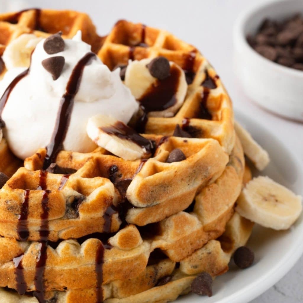 Stack of Chocolate Chip Waffles with whipped cream, banana slices, and chocolate sauce on top. a bowl of chocolate chips in the background.