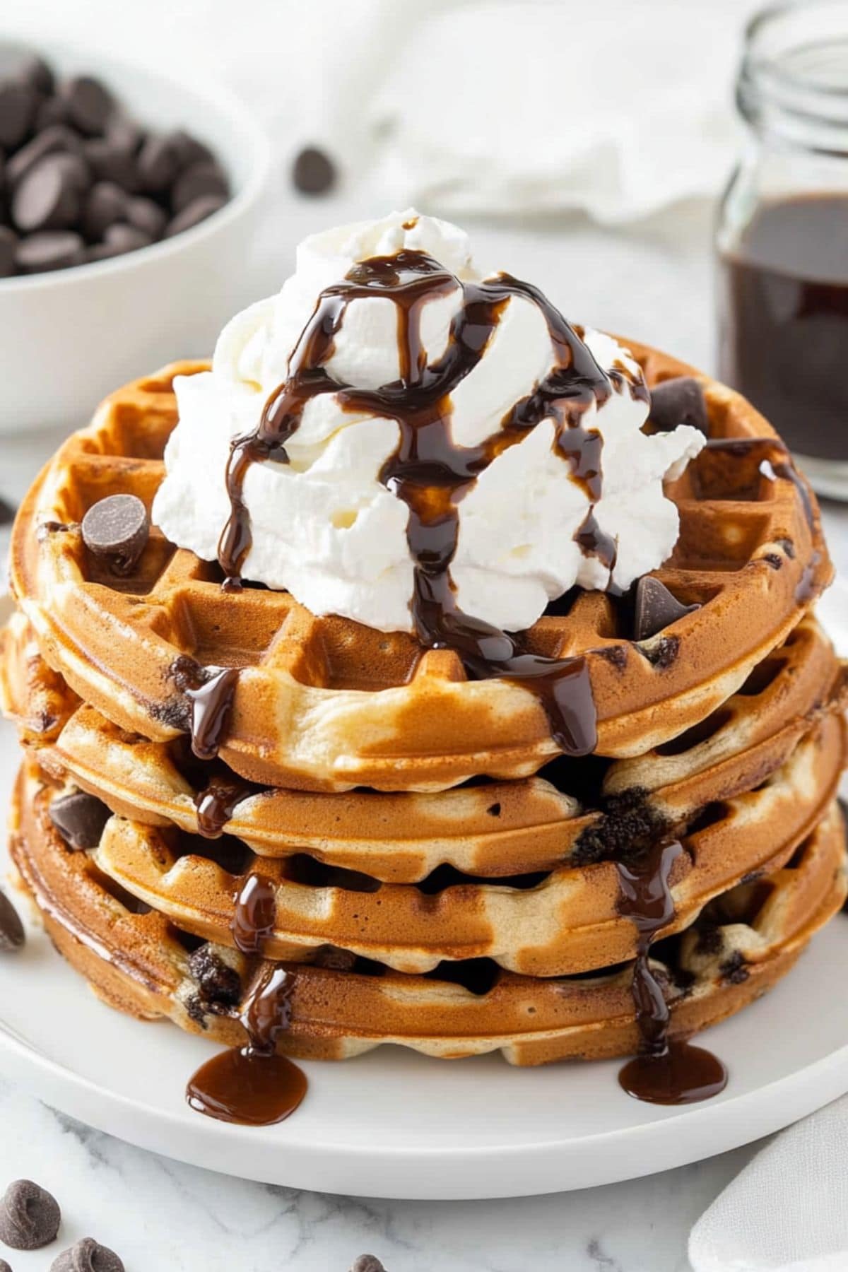 Chocolate chip waffles stacked on a plate, garnished with whipped cream drizzled with chocolate syrup.