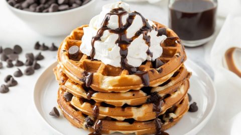 Chocolate chip waffles with whipped cream on top drizzled with chocolate syrup.