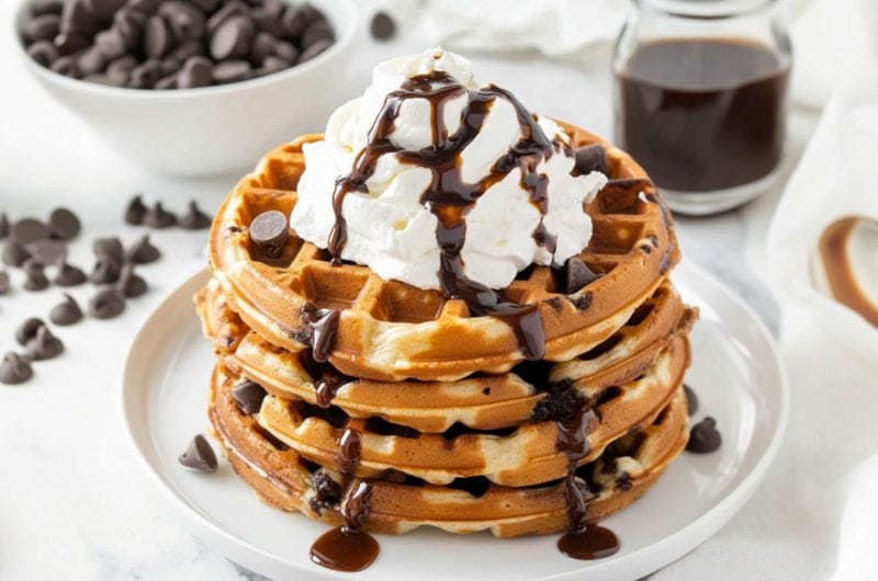 Chocolate chip waffles with whipped cream on top drizzled with chocolate syrup.