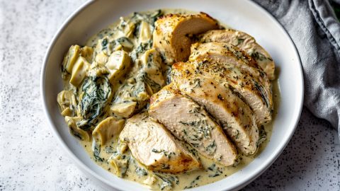 Baked creamy spinach artichoke chicken breast, in a bed of sauce in a white plate.
