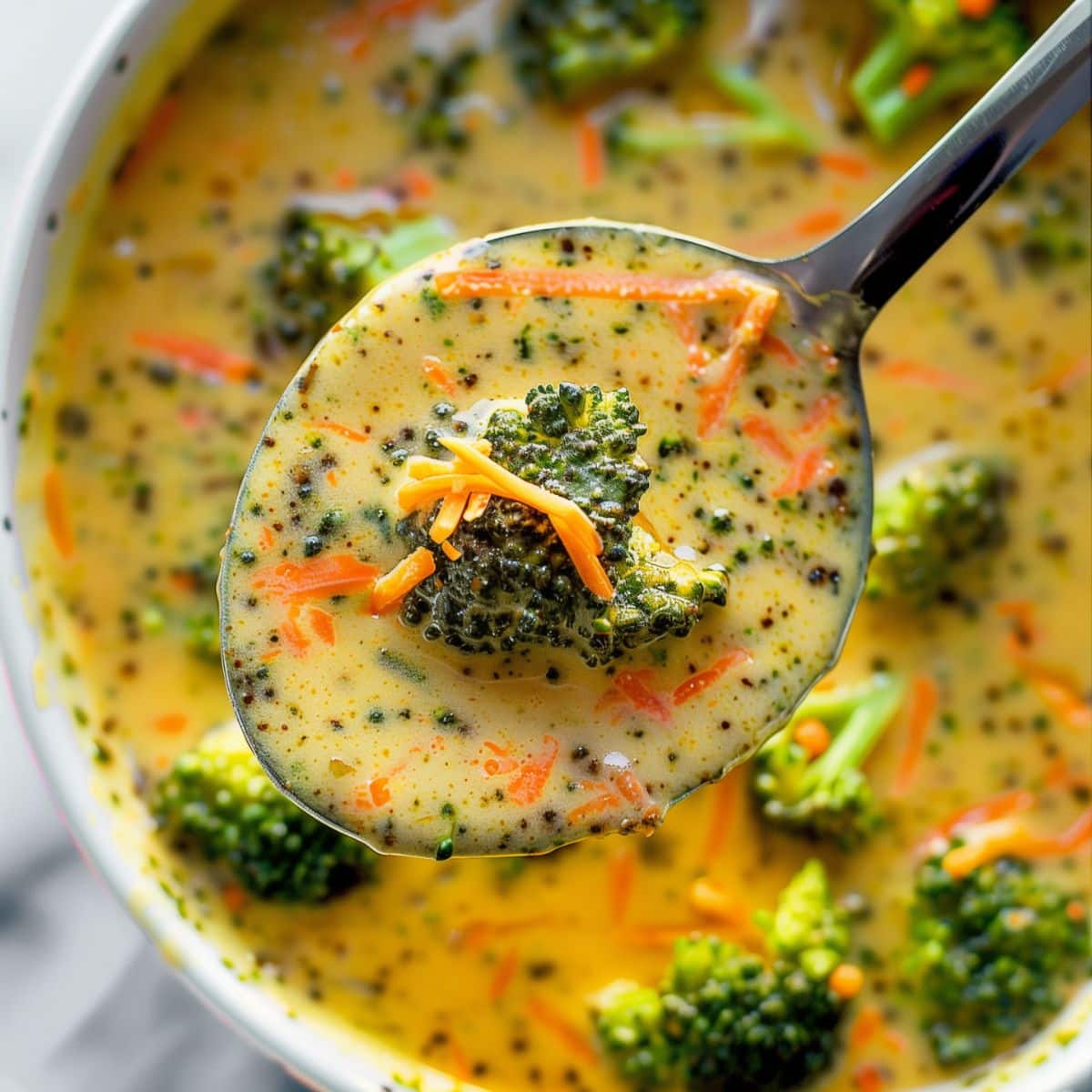 Top View Close Up of Broccoli Cheddar Soup on a Spoon with a Large Broccoli Floret, Shredded Carrots, and Shredded Cheddar Over a Bowl of Broccoli Cheddar Soup