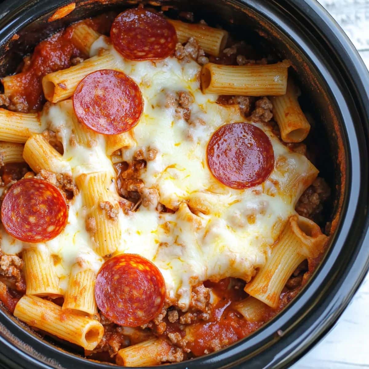 Rigatoni pasta with ground beef, tomato sauce, melted shredded mozzarella cheese, shredded cheddar cheese topped with pepperoni slices and melted cheese cooked inside a black slow cooker pot. 