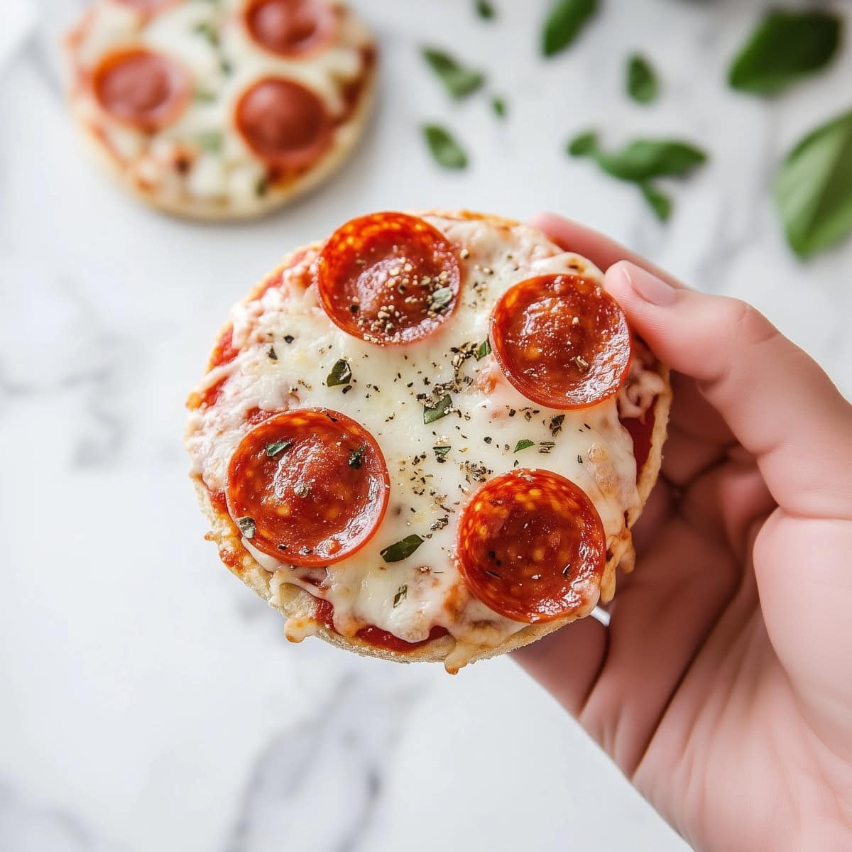 A hand holding English muffin pizza on a white marble background.