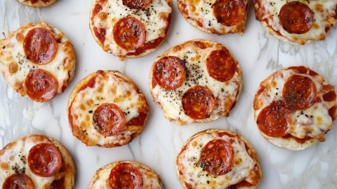 Mini English muffin pizzas on a white marble table topped with melted cheese, pepperoni, and tomato sauce.