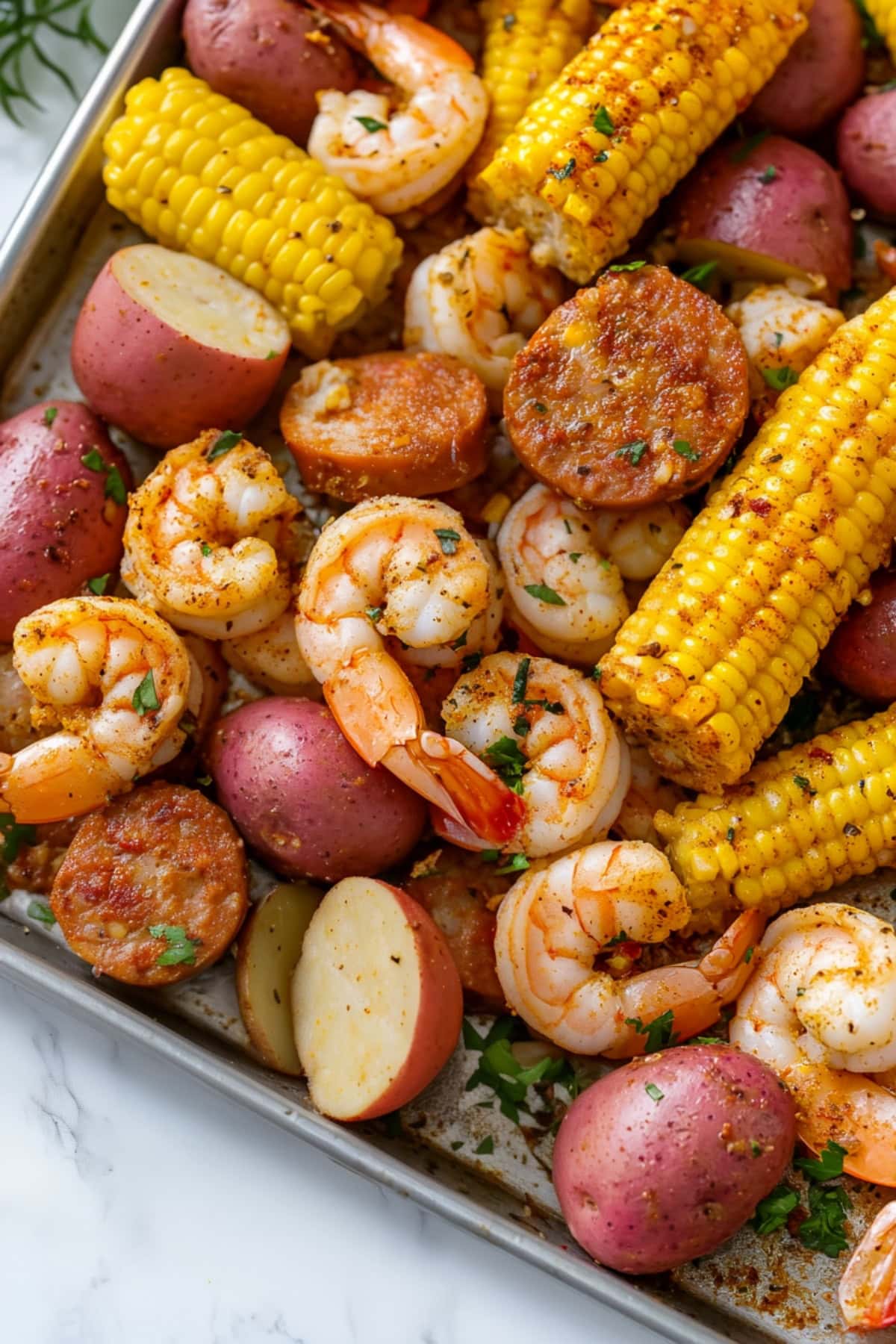 A colorful baking sheet of Frogmore stew, featuring boiled shrimp, corn, andouille sausage, and potatoes, garnished with fresh parsley.