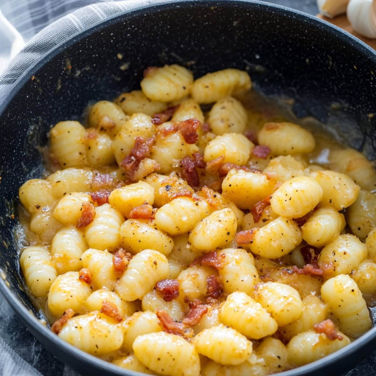 A close-up shot of creamy gnocchi carbonara in a large skillet on a stone table. 