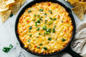 An overhead view of hot corn dip, garnished with herbs and served with chips.