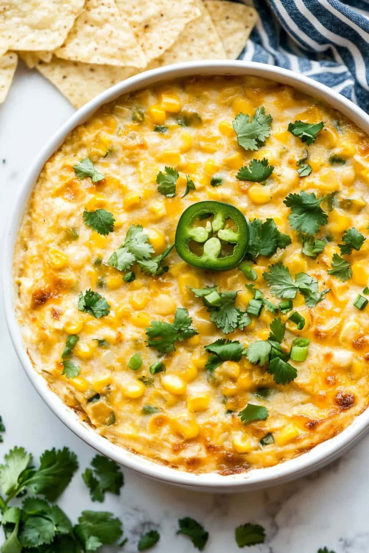 A bowl of hot corn dip garnished with chopped cilantro and jalapeño slices, surrounded by crispy tortilla chips for dipping, top view