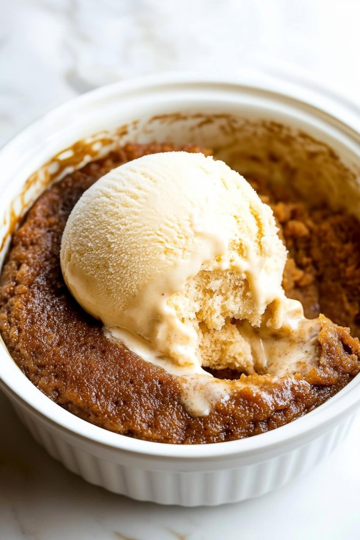 A warm bowl of Indian pudding topped with a scoop of vanilla ice cream, featuring a golden-brown crust and a smooth, rich texture.