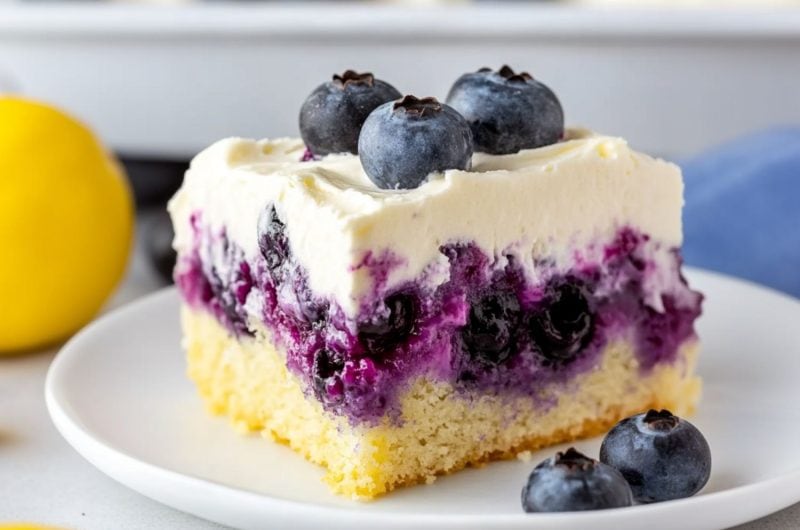 A slice serving of lemon blueberry poke cake garnished with fresh berries served in a white plate.