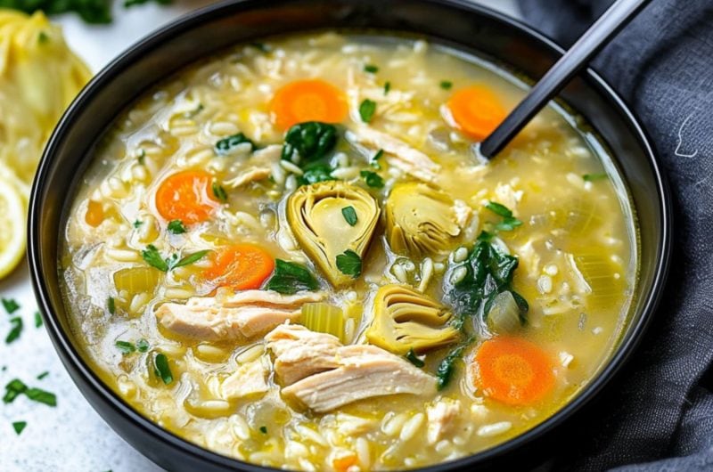 A serving of soup with shredded chicken, artichoke, spinach and orzo pasta served in a black bowl.