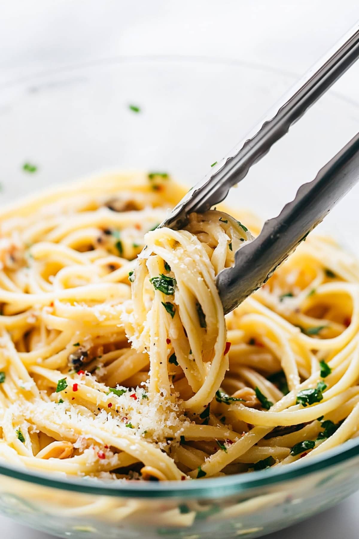 A thong picking up a linguine with clam sauce in a glass bowl.