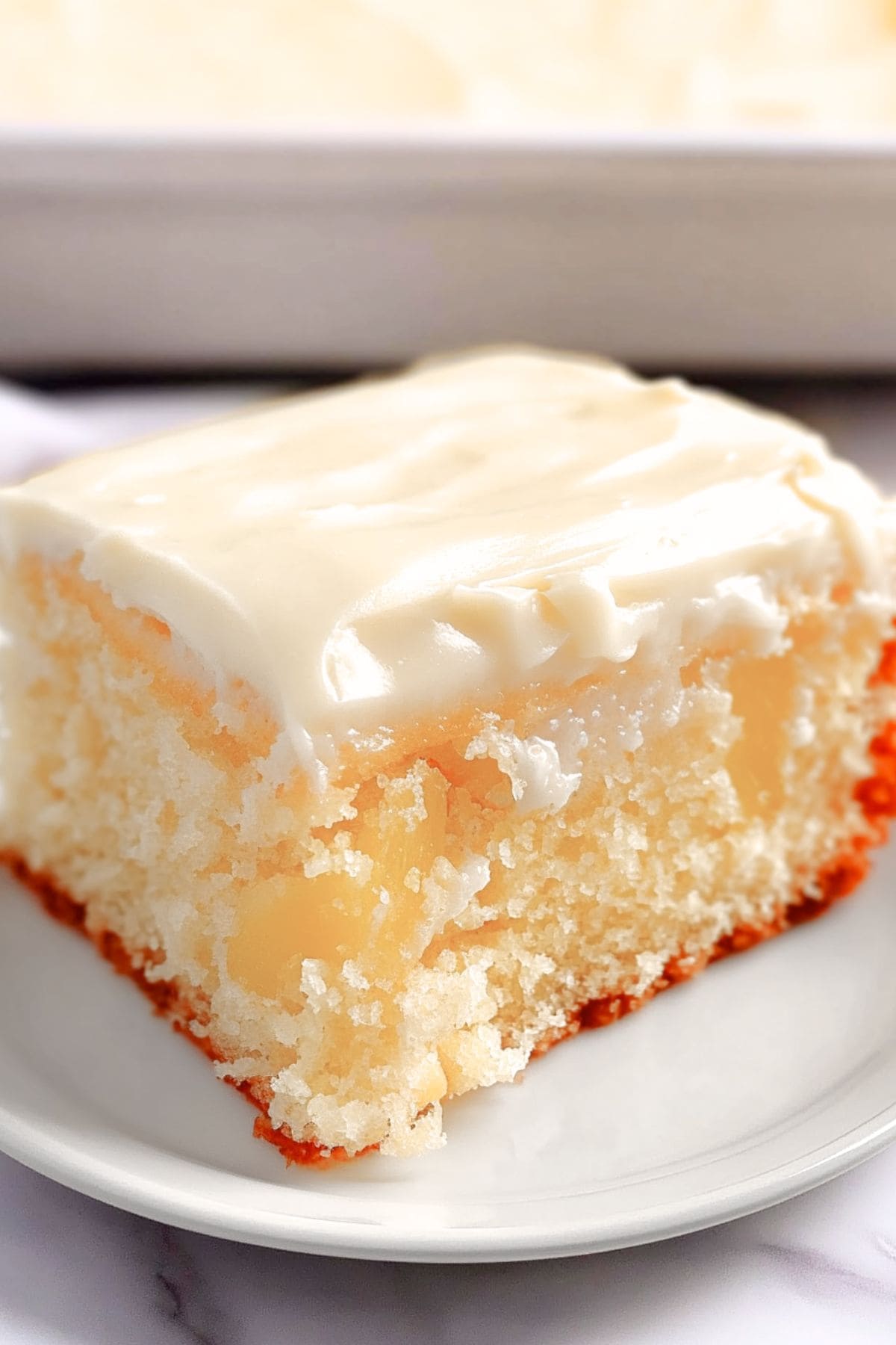 Square slice of moist pineapple cake with cream cheese frosting served on a white plate, close up