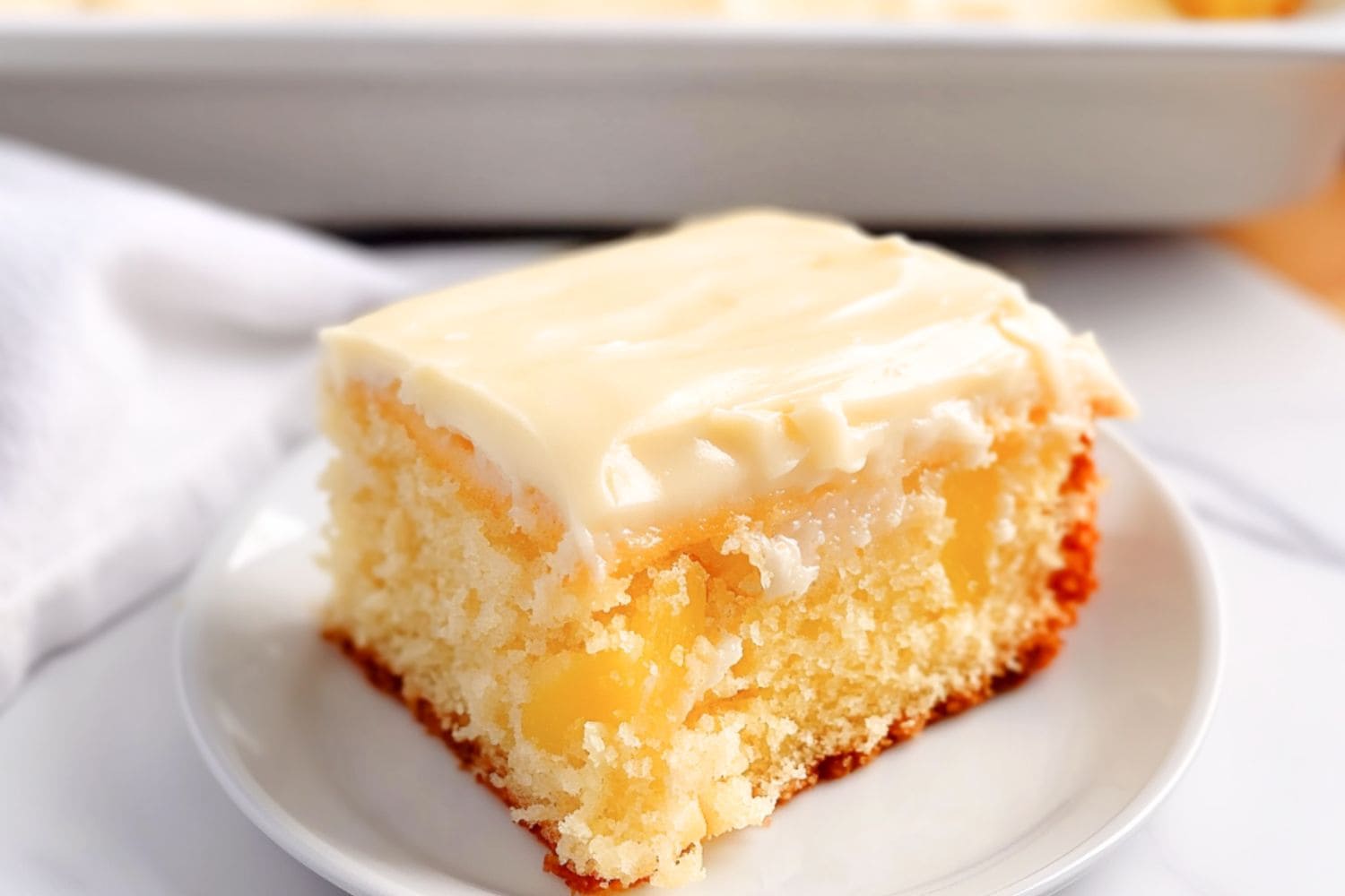 Square slice of moist cake with cream cheese frosting served on a white plate.