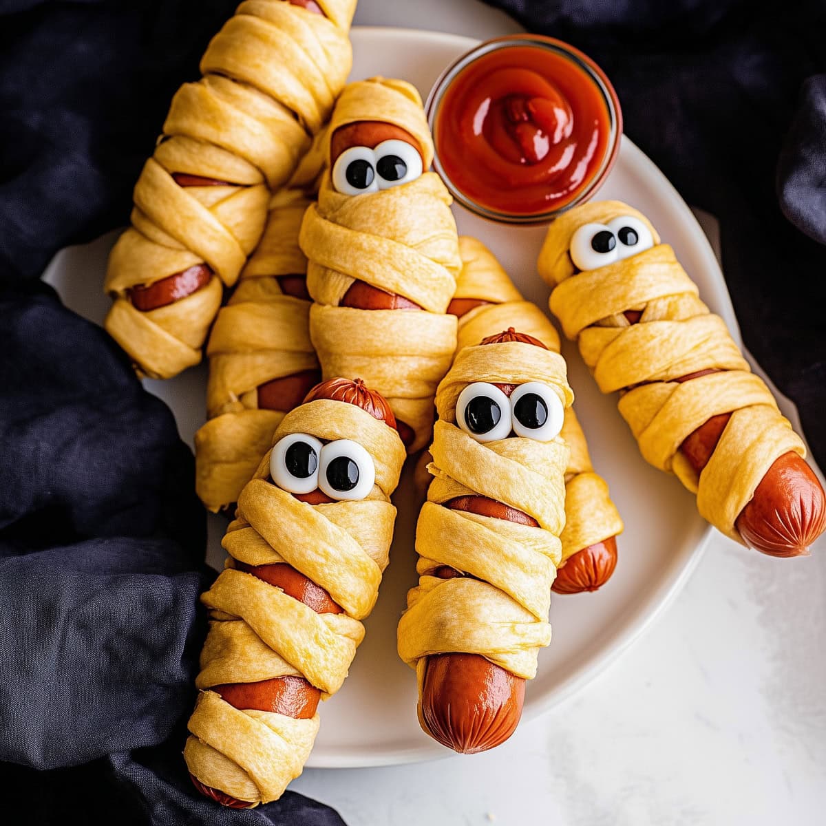 An overhead view of Mummy hot dogs on a plate with ketchup for dipping.