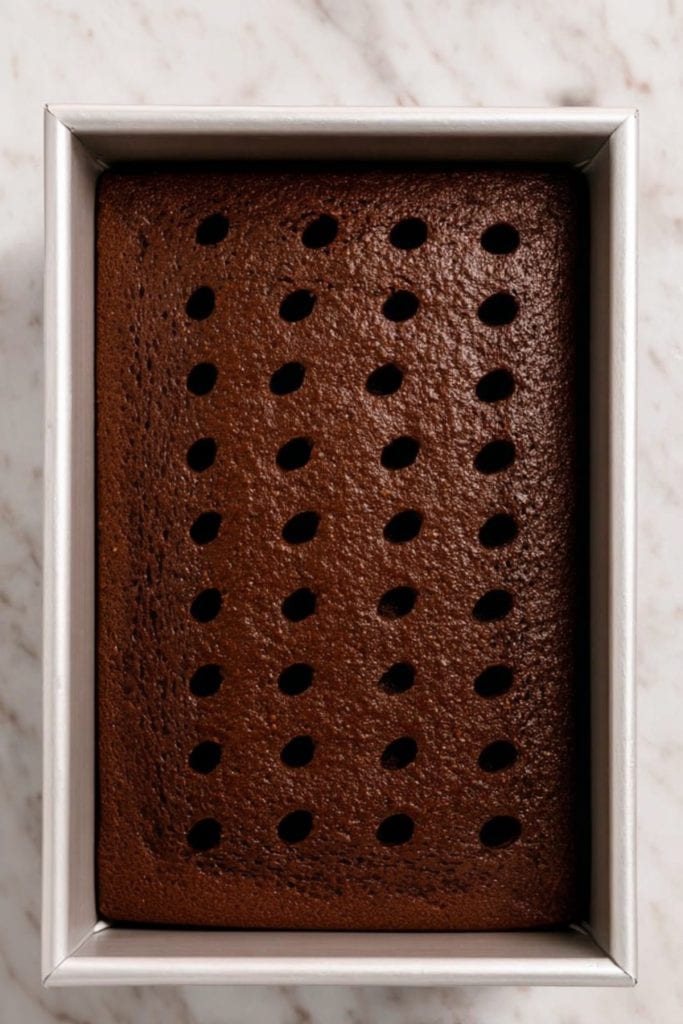 Chocolate cake with poked holes in a stainless pan, top view.