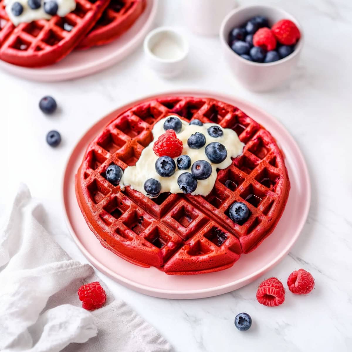 Close-up of fluffy red velvet waffles with cream cheese glaze and fresh berries on a pink plate.