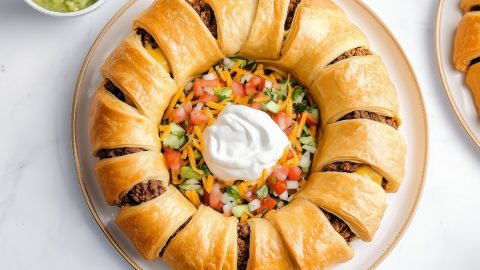 An overhead of taco ring filled with ground beef with colorful veggies and sour cream at the center.