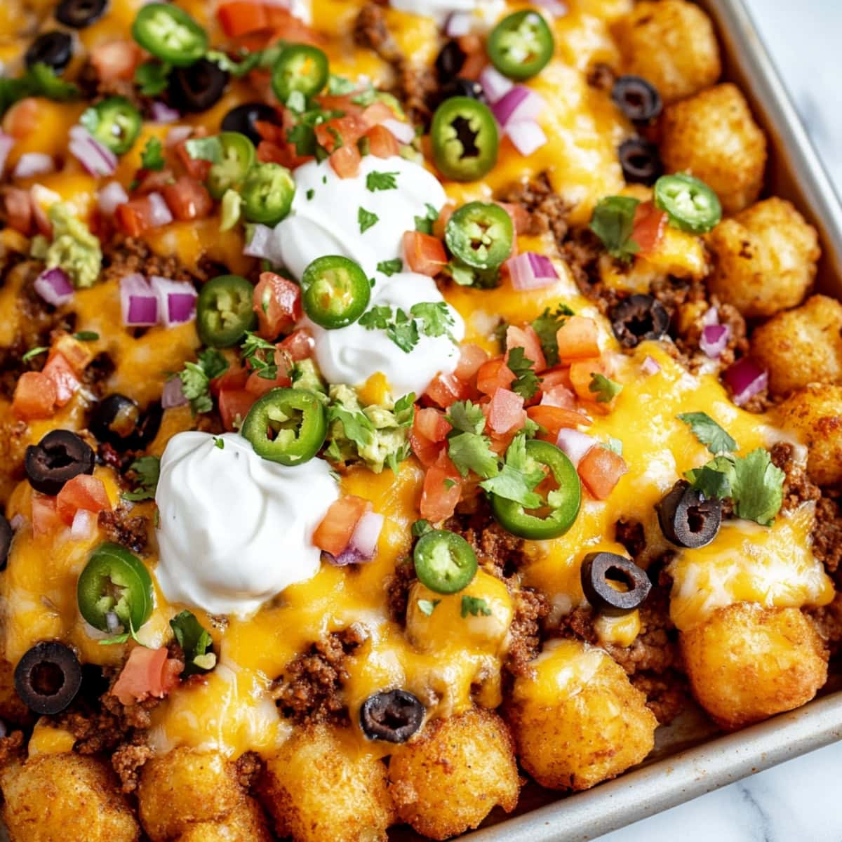 A baking sheet of crispy Totchos topped with melted cheese, jalapeños, sour cream, and red onions.