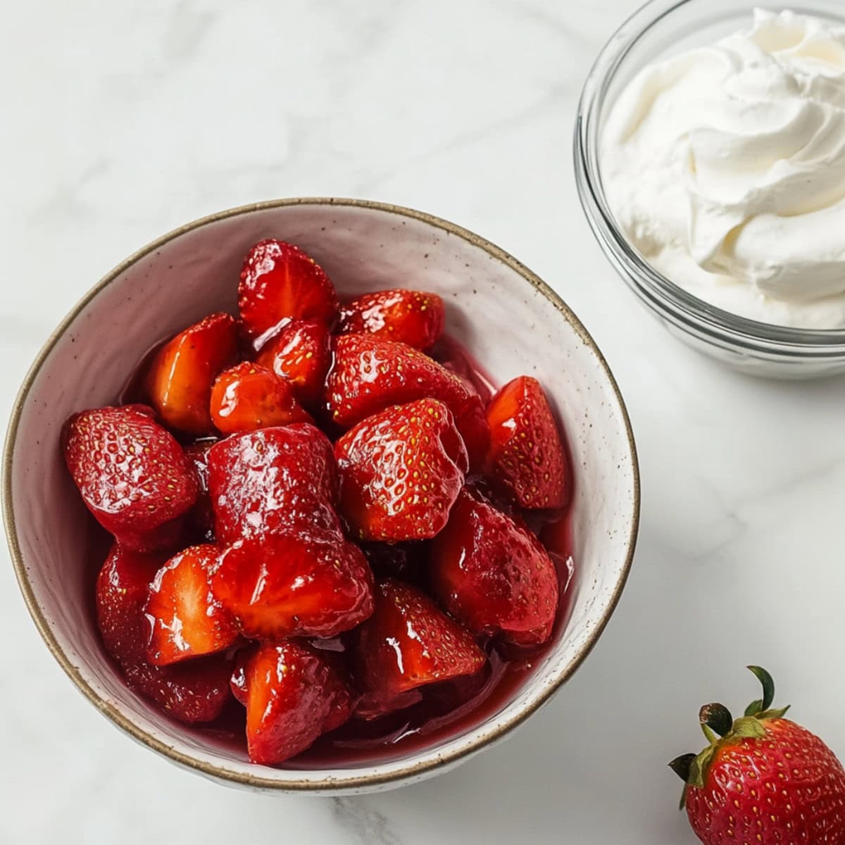 Strawberries coated with sugar syrup served in a white bowl next to a bowl with whipped cream.