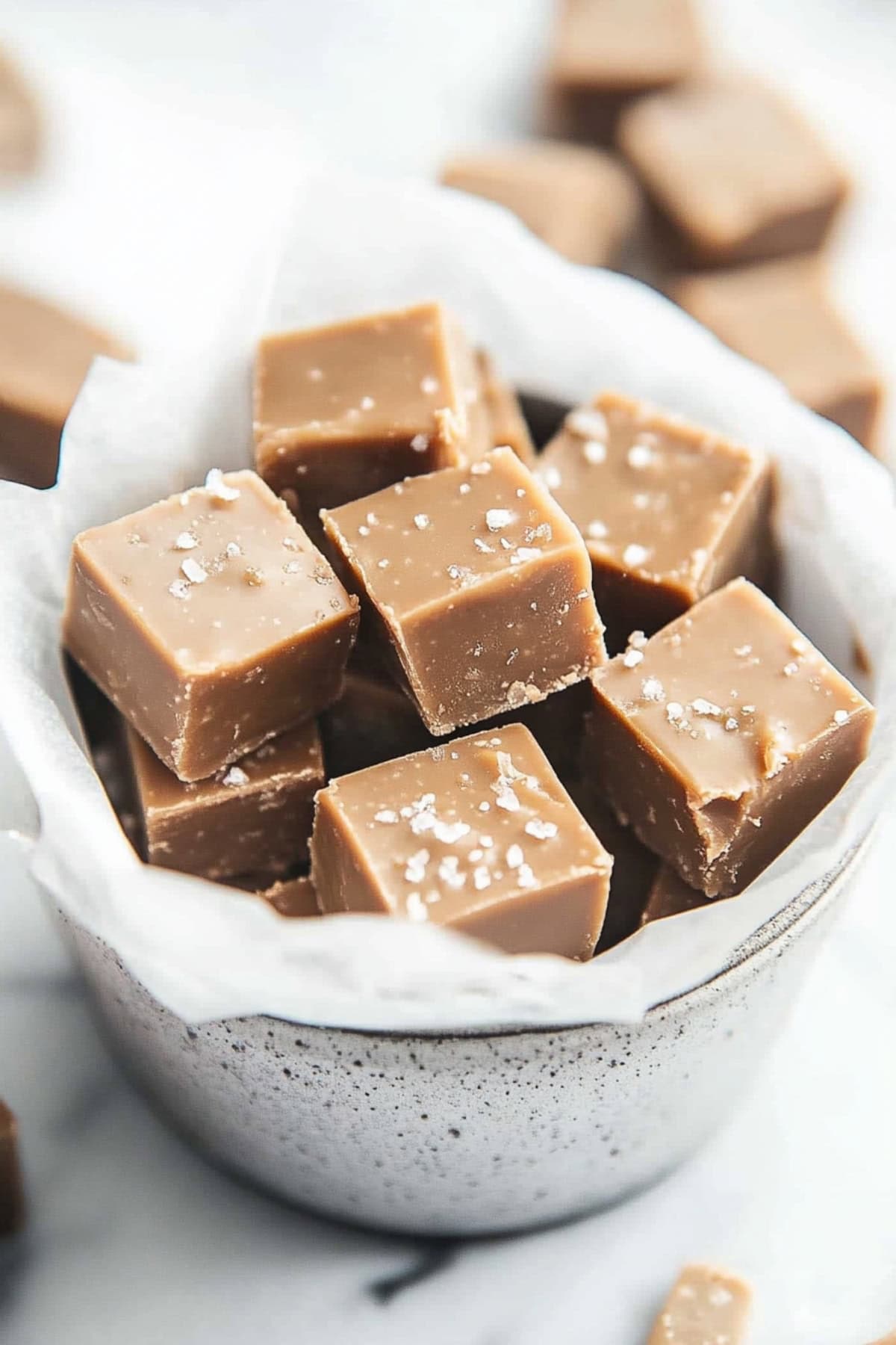 Salted caramel fudge squares inside a bowl with parchment paper on a white marble table. 