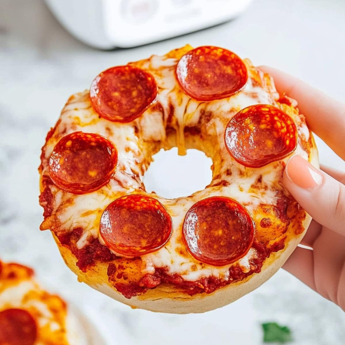 A hand holding a piece of cheesy bagel pizza with pepperoni.