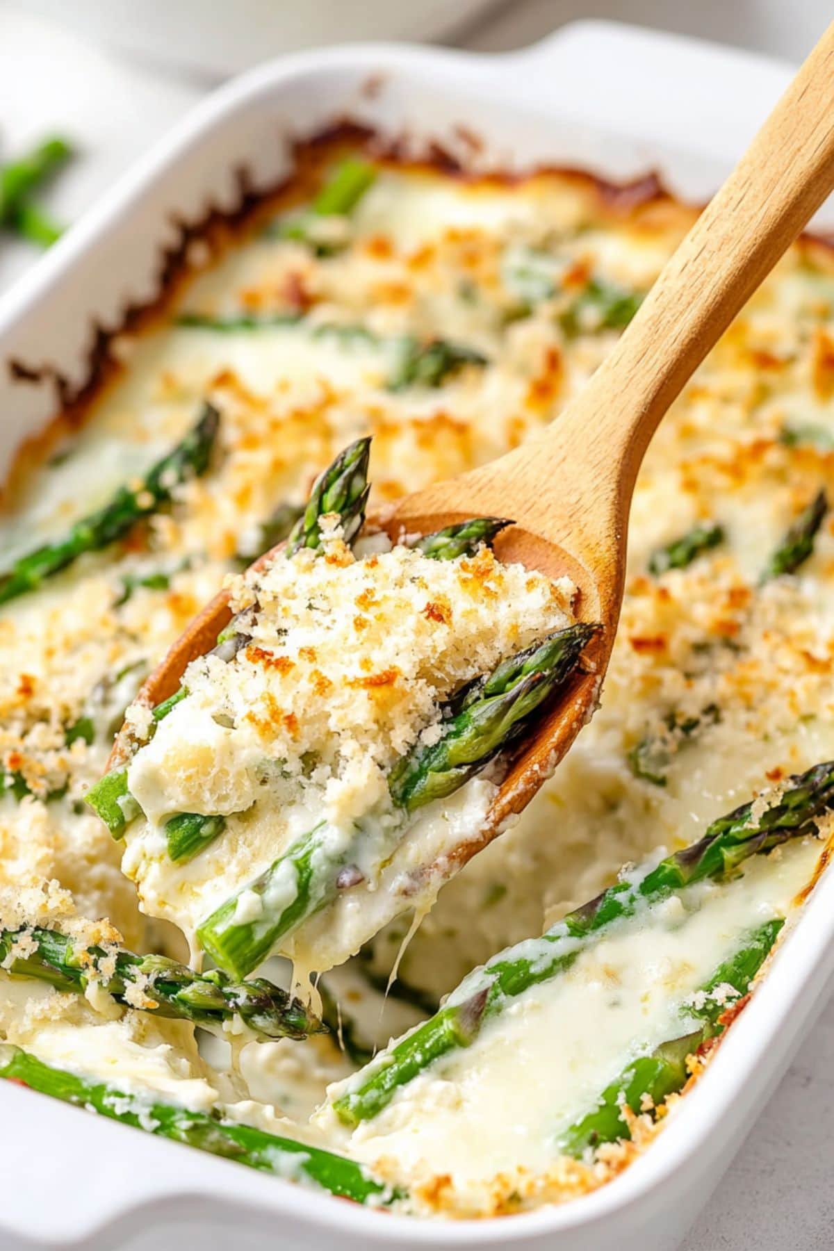 Wooden ladle lifting a serving of creamy asparagus casserole from baking dish.