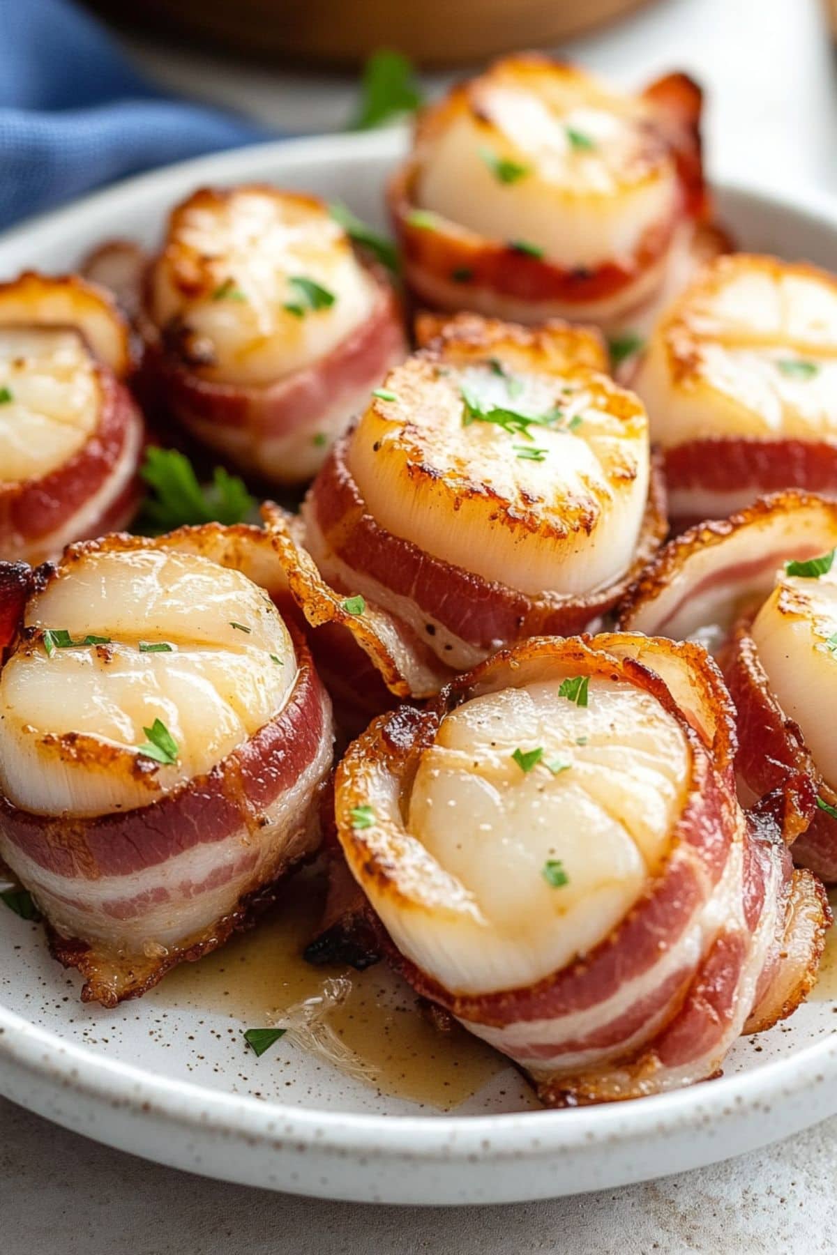 Bunch of bacon wrapped scallops in a white plate garnished with chopped parsley.