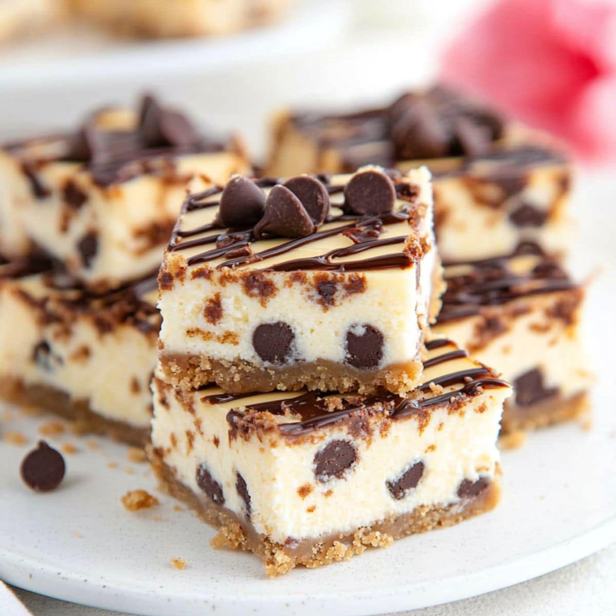 Square slices of cannoli cheesecake bars stacked on a white plate drizzled with chocolate syrup.