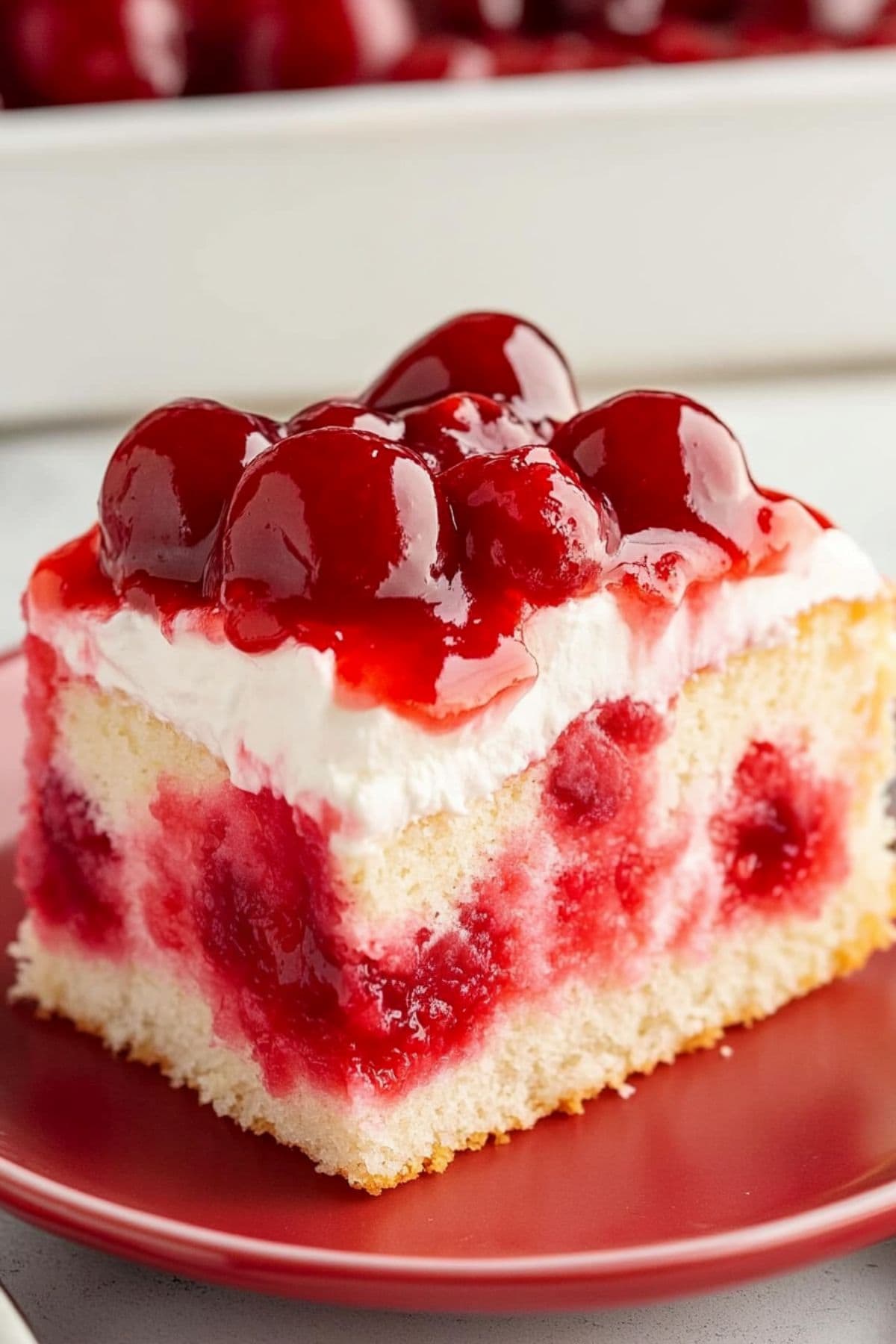 Square slice of vanilla cake with Jello streaks, topped with whipped cream and cherry pie filling served on a red plate.