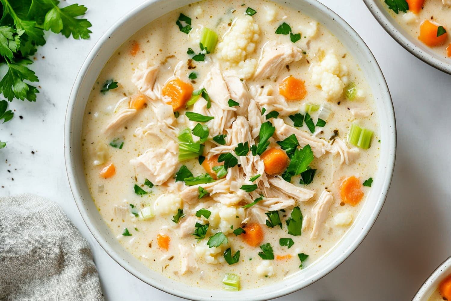 A warm, comforting soup with creamy base, pieces of chicken, soft cauliflower, diced carrots and celery on a white marble table.