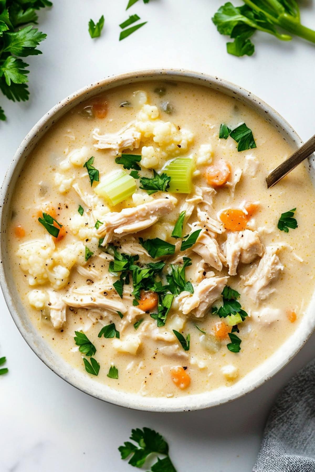 Large bowl of chicken cauliflower soup with celery stalks and carrots, garnished with parsley, top view