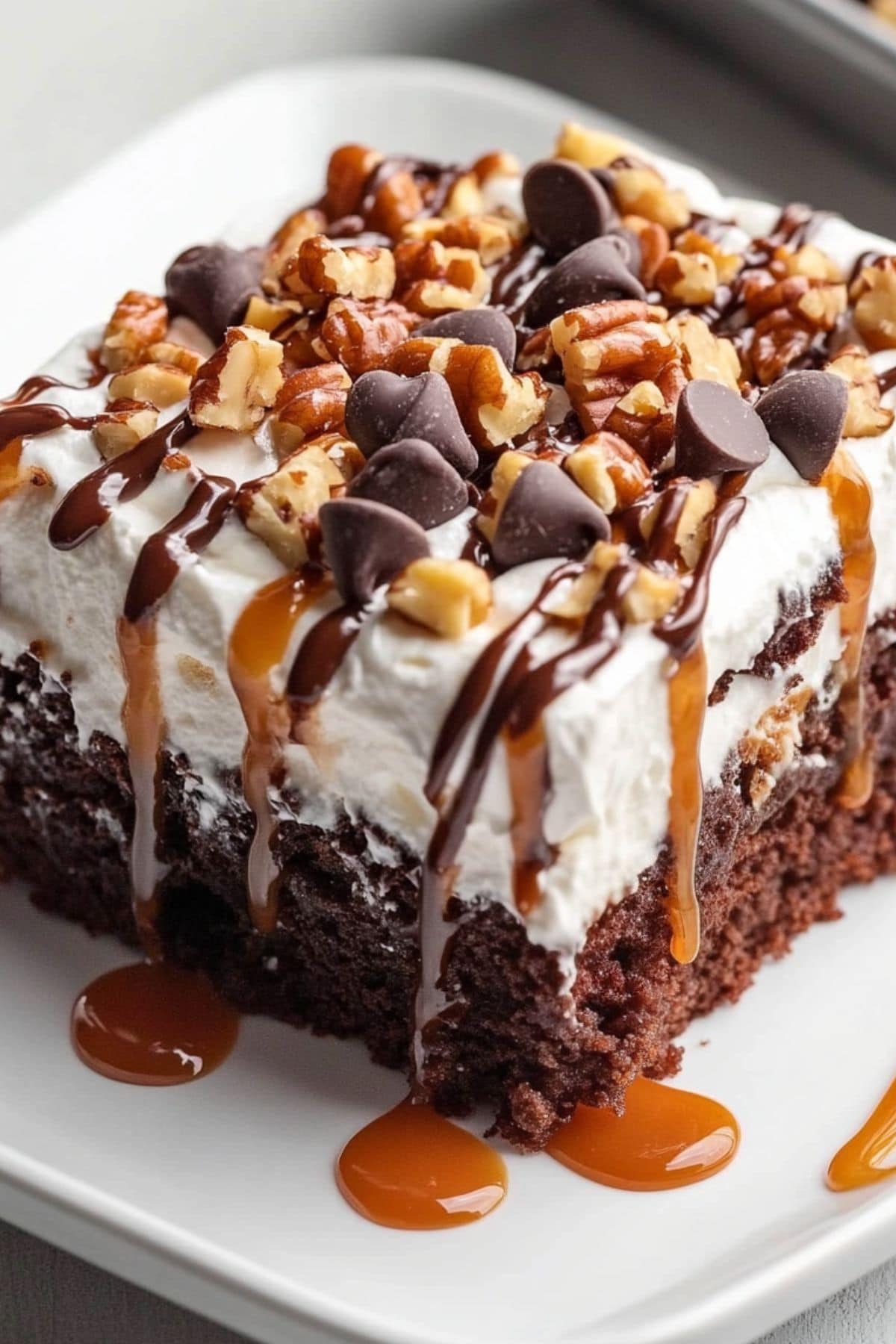 Fudgy chocolate poke cake with whipped cream, chopped pecans, and chocolate chips on top. It's drizzled with chocolate and caramel sauce. 