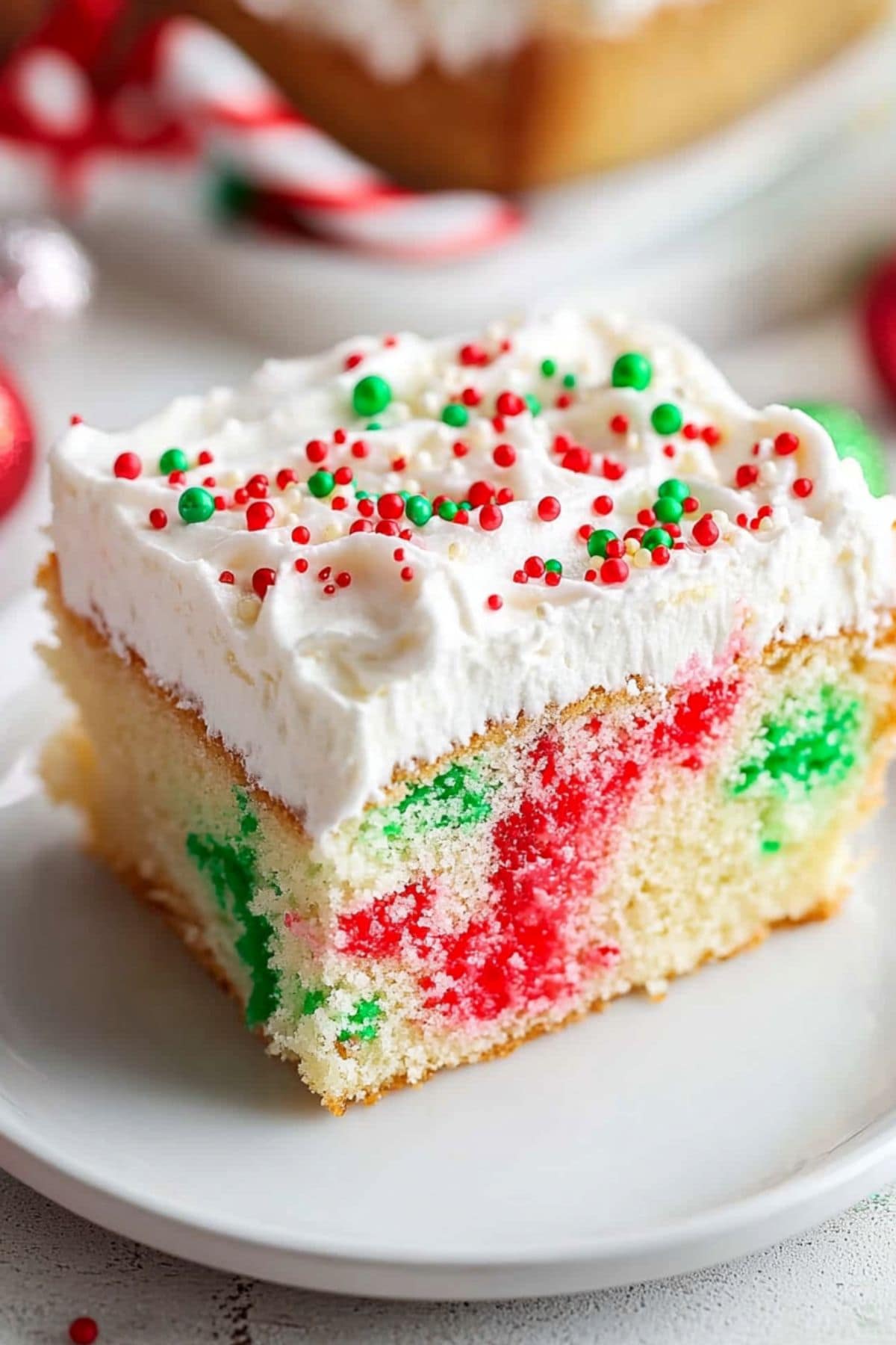 Square slice of Christmas poke cake with red and green Jello O streaks, topped with a layer of whipped cream garnished with candy sprinkles.