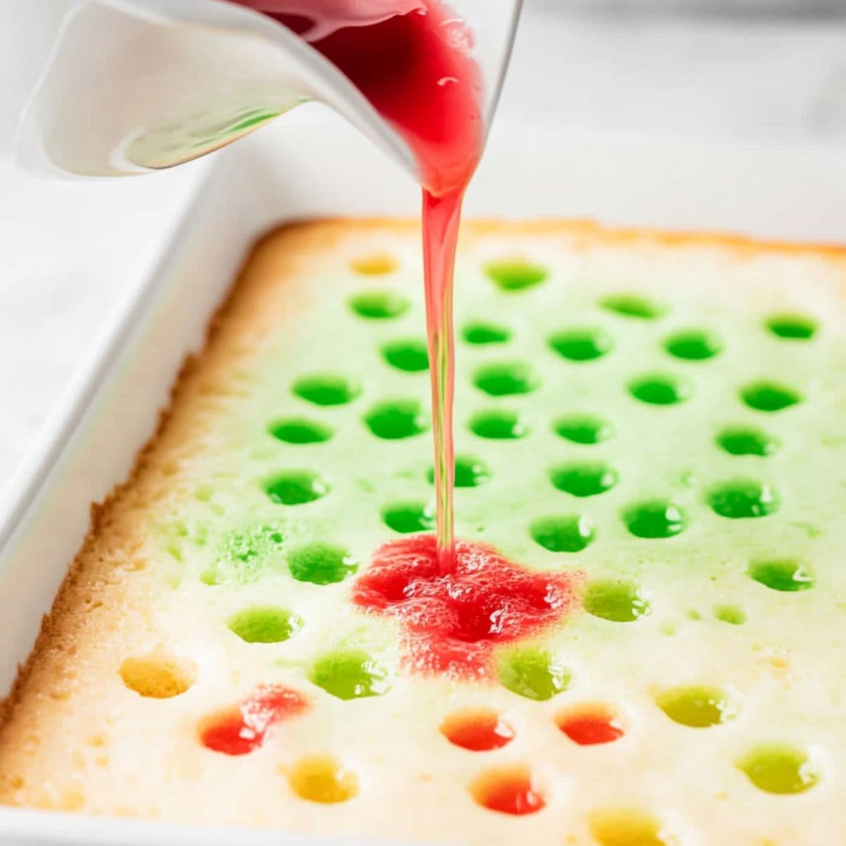 Red jello in a bowl poured over cake with holes in a rectangular baking dish.