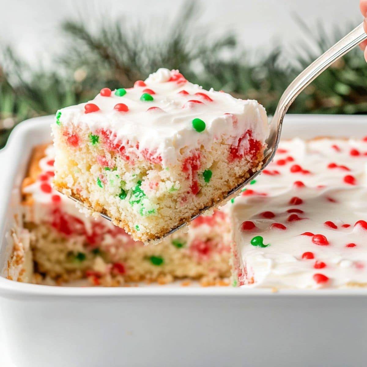 Cake ladle lifting a square slice of red and green infused poke cake with a layer of whipped cream with candy sprinkles.