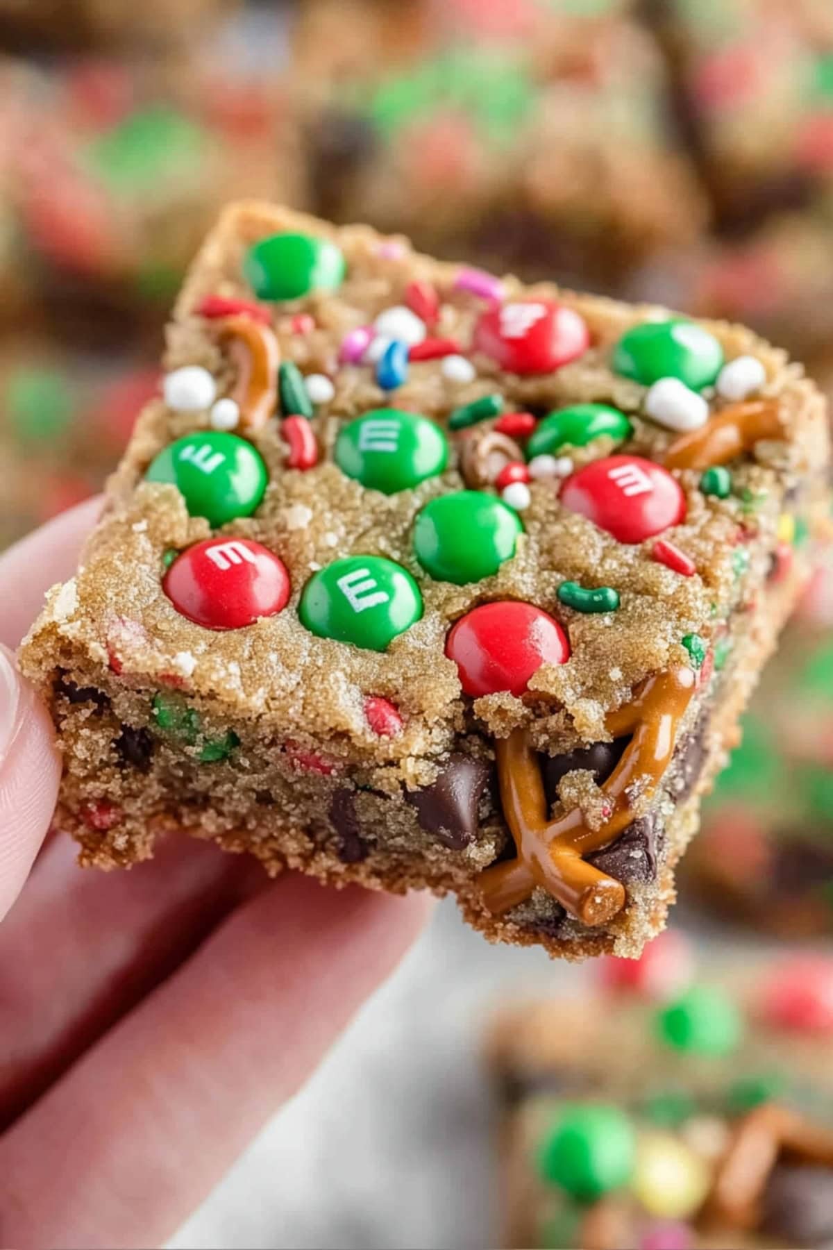 A hand holding a piece of Christmas cookie bar with chocolate chips, M&M candies and crushed pretzels.