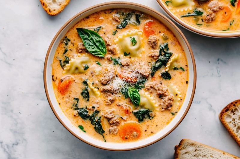 Two bowls of creamy ravioli soup featuring baby spinach, carrots and Italian sausage.