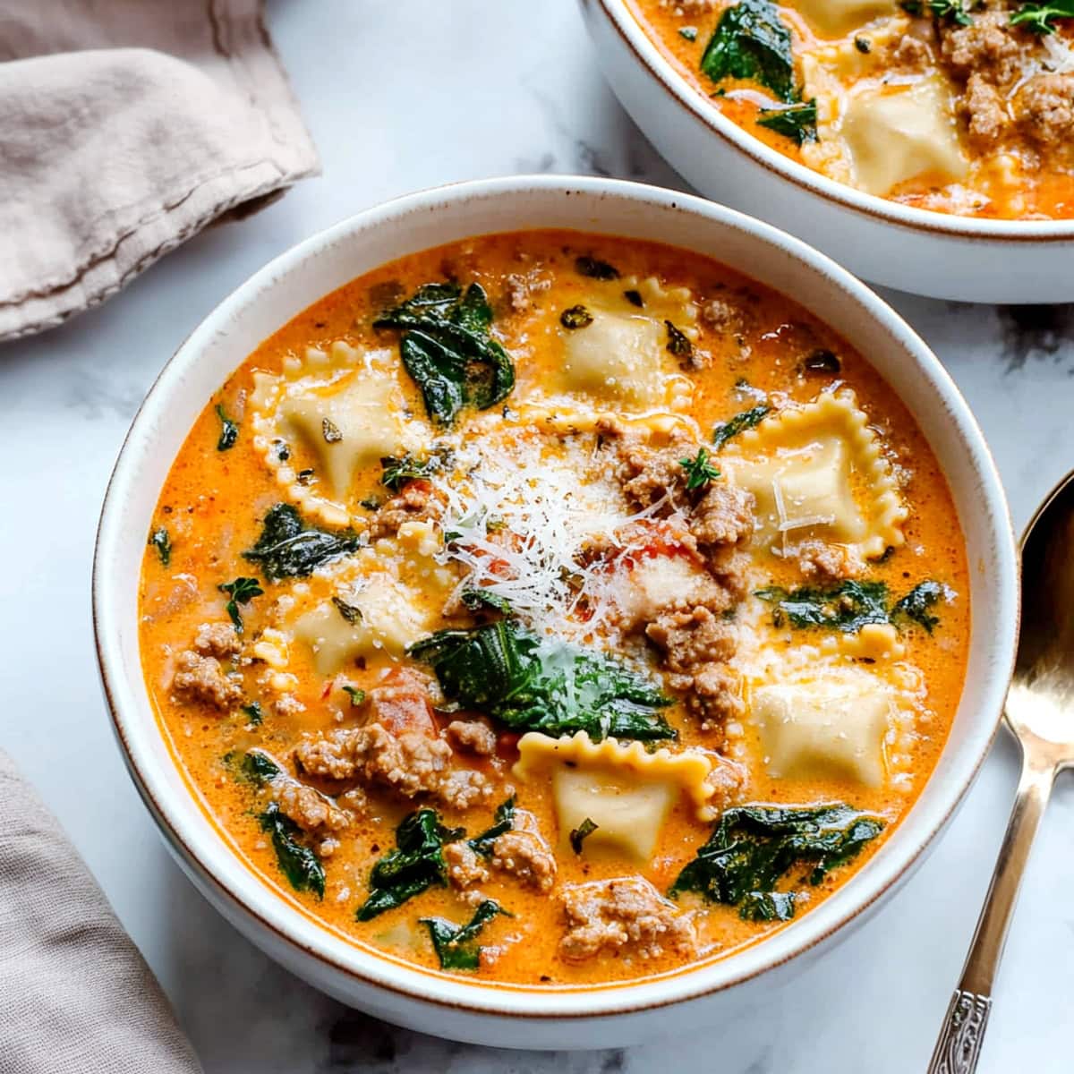 Two bowls of creamy ravioli soup with baby spinach and ground meat.