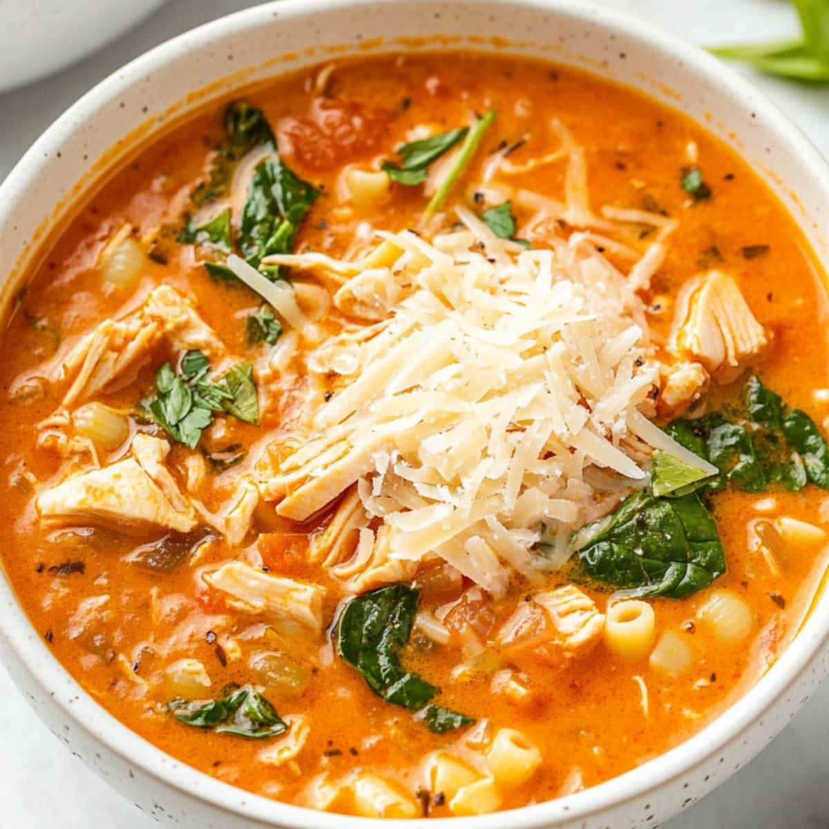 Cheesy chicken parmesan soup with tomato base broth, shredded chicken, pasta and chopped baby spinach served on a white bowl.