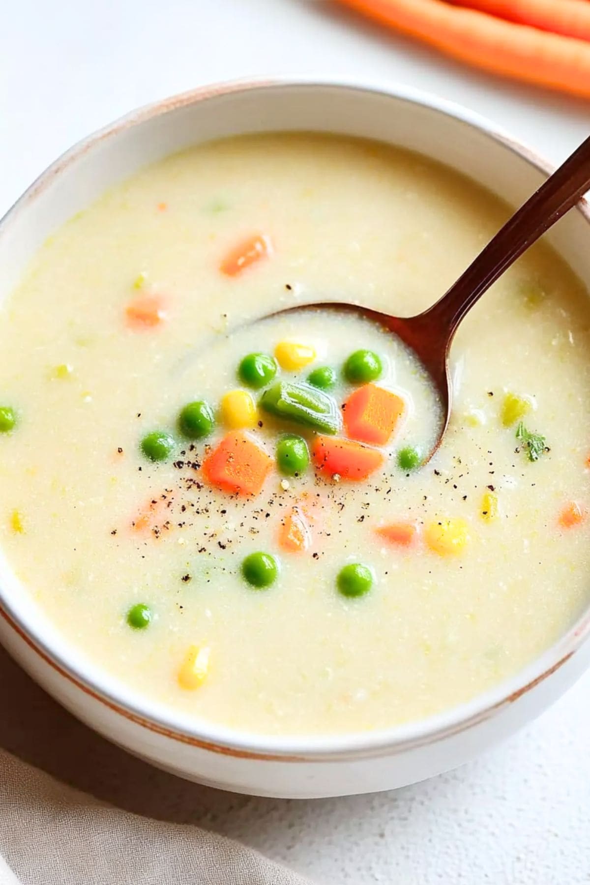 Spoon scooping serving of creamy soup with mixed veggies from a white bowl.