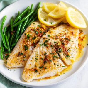 Oven baked tilapia fillet served in a white plate with lemon wedge and steamed beans.